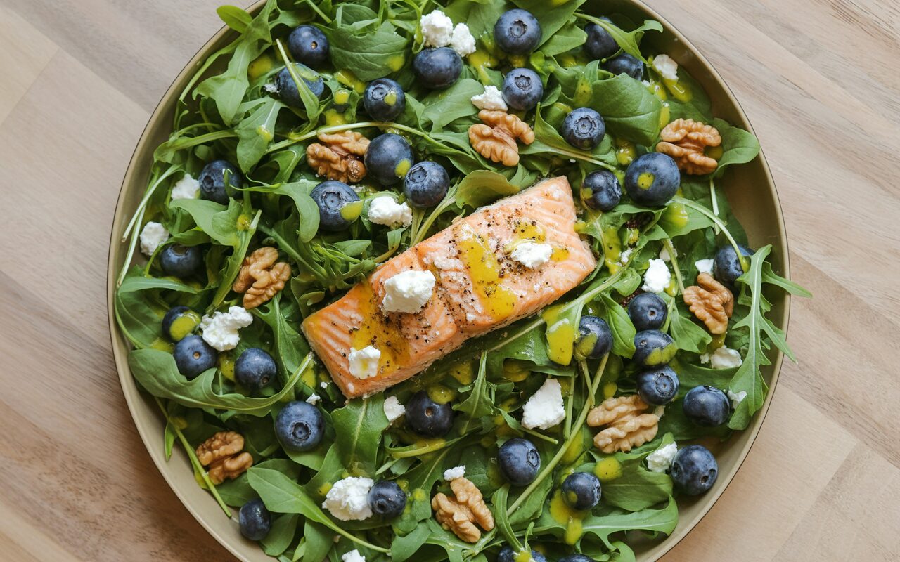 A bright, overhead flat lay of a salmon and blueberry arugula salad in a wide shallow bowl on a light wood surface, with crumbled goat cheese, walnuts, and fresh blueberries scattered throughout, drizzled with lemon vinaigrette, natural afternoon light, food photography style, vibrant and fresh